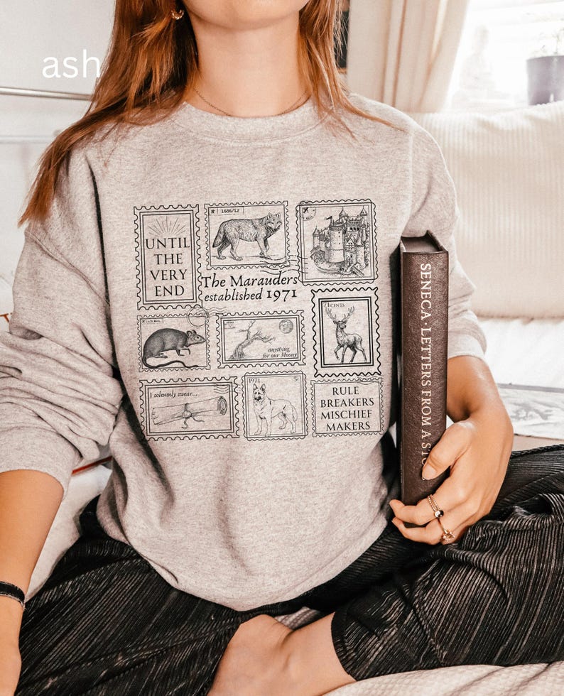 K&ouml;nnte beinhalten: Ein graues Sweatshirt mit einem grafischen Druck von Briefmarken mit verschiedenen Bildern, darunter ein Wolf, ein Schloss, eine Ratte, ein Baum, ein Hirsch, eine Schriftrolle und ein Hund. Der Text auf den Briefmarken lautet "Until the very end", "The Marauders established 1971" und "Rule Breakers Mischief Makers".