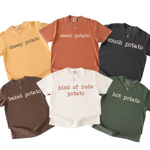 May include: Six short-sleeved t-shirts in shades of yellow, orange, black, brown, white, and green. Each shirt displays a different potato-related phrase in white text, like "cheesy potato" and "sweet potato".