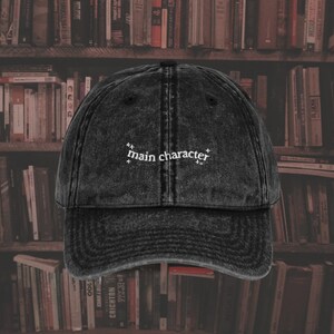 Könnte beinhalten: Eine schwarze Baseballkappe aus Denim mit dem Schriftzug "main character" in weiß auf der Vorderseite.