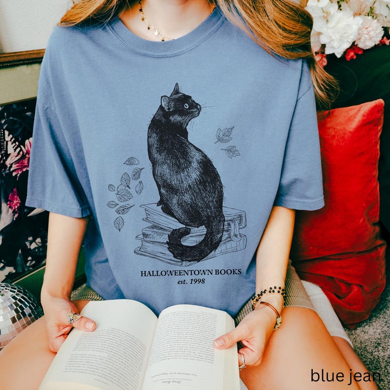 Op de afbeelding: Lichtblauw T-shirt met een illustratie van een zwarte kat die op een stapel boeken en bladeren zit. De tekst "Halloweentown Books est. 1998" staat eronder. Het shirt is gemaakt van zacht materiaal.