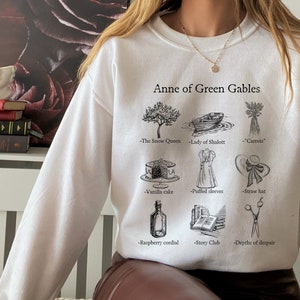 Op de afbeelding: Wit sweatshirt met een zwarte grafische print van illustraties en tekst die "Anne of Green Gables" zegt. De illustraties omvatten een boom, een boot, wortels, een taart, een jurk, een hoed, een fles, een stapel boeken en een schaar.