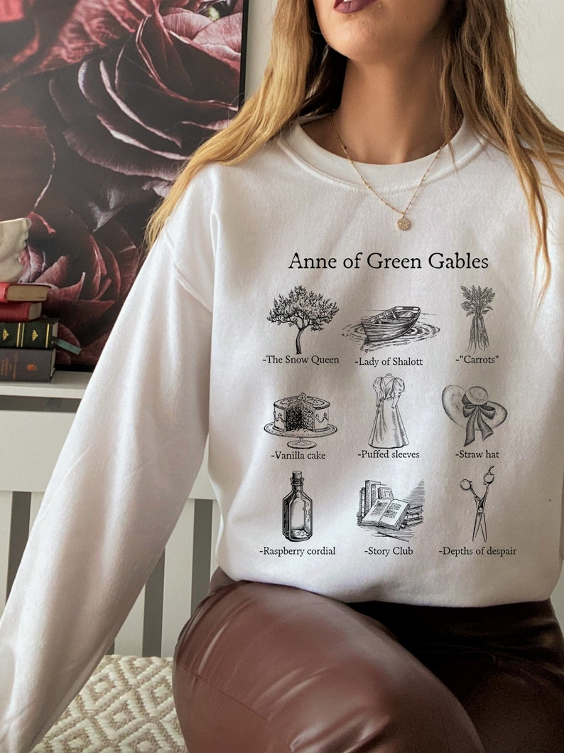 Op de afbeelding: Wit sweatshirt met een zwarte grafische print van illustraties en tekst die "Anne of Green Gables" zegt. De illustraties omvatten een boom, een boot, wortels, een taart, een jurk, een hoed, een fles, een stapel boeken en een schaar.