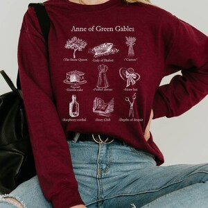 Op de afbeelding: Een bordeauxrood sweatshirt met een witte grafische print van illustraties en tekst die "Anne of Green Gables" zegt. De illustraties omvatten een boom, een boot, wortels, een taart, een jurk, een hoed, een fles, een stapel boeken en een schaar.