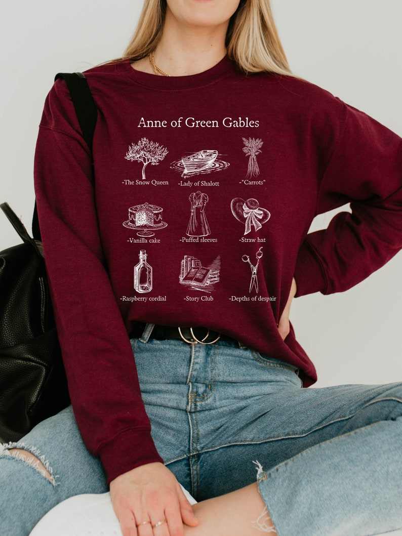 Op de afbeelding: Een bordeauxrood sweatshirt met een witte grafische print van illustraties en tekst die "Anne of Green Gables" zegt. De illustraties omvatten een boom, een boot, wortels, een taart, een jurk, een hoed, een fles, een stapel boeken en een schaar.