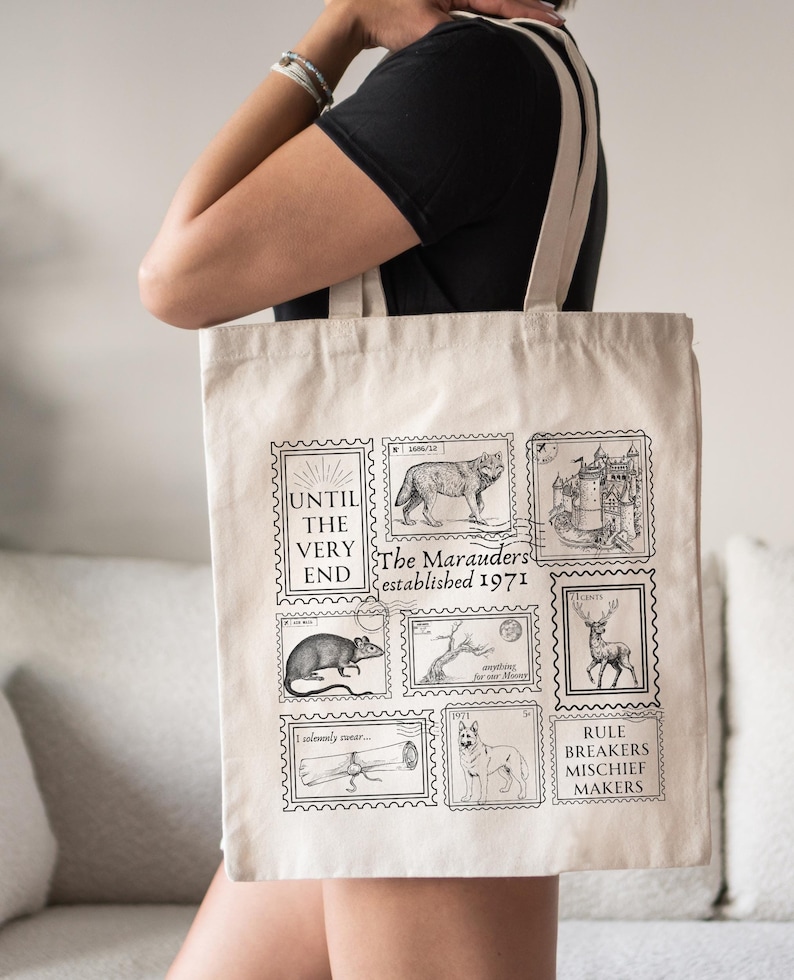 Puede incluir: Una bolsa de lona blanca con ilustraciones negras de un lobo, un castillo, una rata, un &aacute;rbol, un ciervo y un perro. La bolsa tiene el texto "The Marauders established 1971" y "Until the very end".