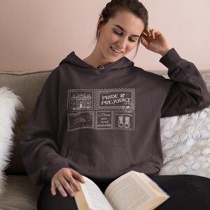 May include: A dark brown hoodie featuring a graphic design inspired by "Pride & Prejudice." The design includes the title and illustrations resembling postage stamps. The hoodie has a front pocket and a drawstring hood.