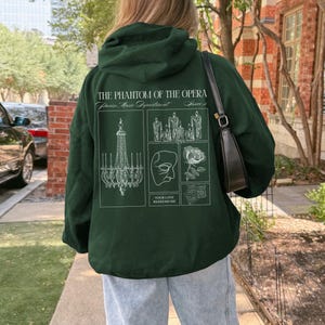 May include: Dark green hoodie with a white graphic design from "The Phantom of the Opera." The design includes a chandelier, candles, a mask, a rose, and the text "Your Love Redeems Me."