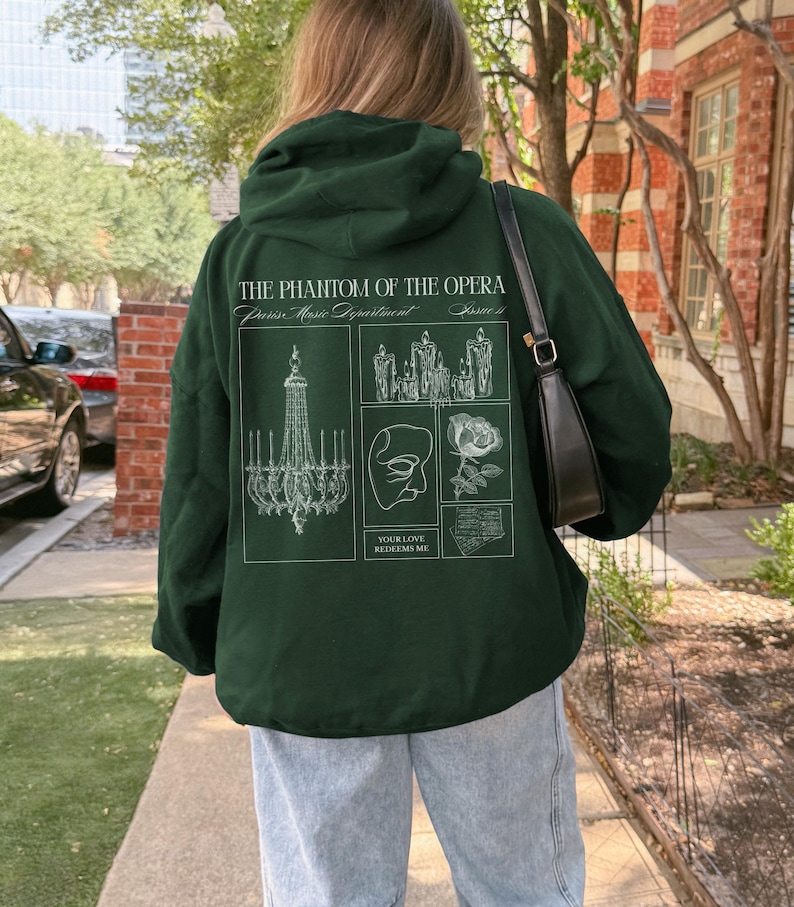 May include: Dark green hoodie with a white graphic design from "The Phantom of the Opera." The design includes a chandelier, candles, a mask, a rose, and the text "Your Love Redeems Me."