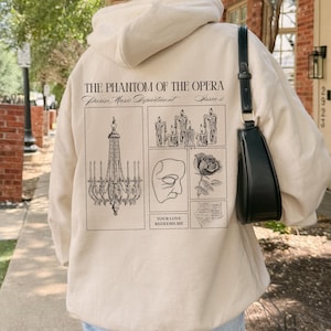 May include: Beige hoodie featuring a graphic design from "The Phantom of the Opera." The design includes a chandelier, candles, a mask, a rose, and the text "YOUR LOVE REDEEMS ME."