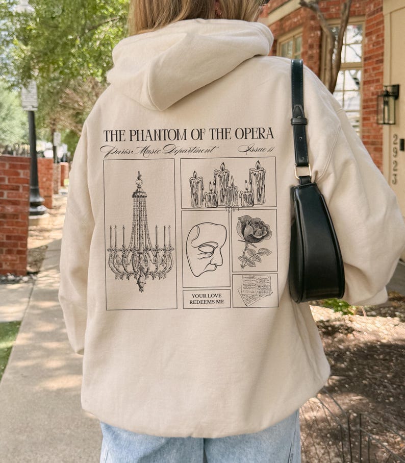 May include: Beige hoodie featuring a graphic design from "The Phantom of the Opera." The design includes a chandelier, candles, a mask, a rose, and the text "YOUR LOVE REDEEMS ME."