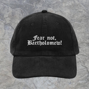 May include: A black corduroy baseball cap featuring the white embroidered text Fear not, Bartholomew! centered on the front panel. The cap has a structured crown, a curved brim, and a button at the top, showcasing a classic six-panel design.