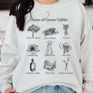 Op de afbeelding: Een grijs sweatshirt met de tekst "Anne of Green Gables" en illustraties van verschillende voorwerpen uit het boek, waaronder een boom, een boot, wortels, een taart, een jurk, een hoed, een fles, een stapel boeken en een schaar.