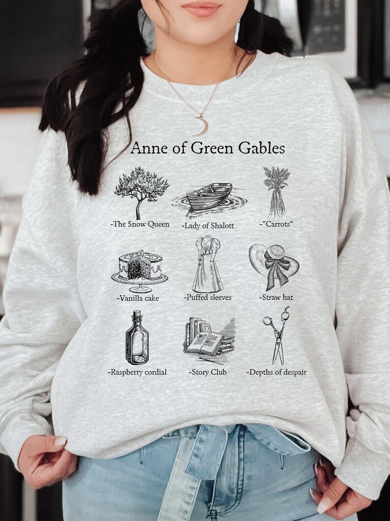 Op de afbeelding: Een grijs sweatshirt met de tekst "Anne of Green Gables" en illustraties van verschillende voorwerpen uit het boek, waaronder een boom, een boot, wortels, een taart, een jurk, een hoed, een fles, een stapel boeken en een schaar.