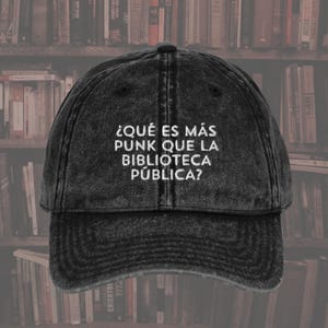 May include: A black, distressed baseball cap with the text "¿QUÉ ES MÁS PUNK QUE LA BIBLIOTECA PÚBLICA?" embroidered in white. The cap has a curved brim and an adjustable strap. The background shows a blurred image of bookshelves.