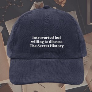 May include: A navy blue corduroy baseball cap with white embroidered text that reads "introverted but willing to discuss The Secret History".