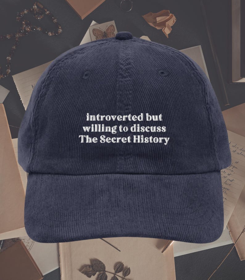 May include: A navy blue corduroy baseball cap with white embroidered text that reads "introverted but willing to discuss The Secret History".