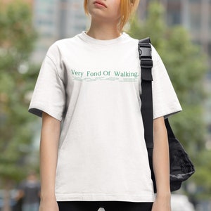 May include: A white t-shirt with the text "Very Fond Of Walking." printed in green on the front. The person wearing the shirt is standing in an urban setting.