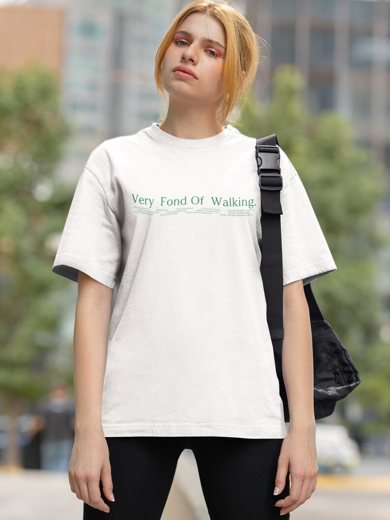 May include: A white t-shirt with the text "Very Fond Of Walking." printed in green on the front. The person wearing the shirt is standing in an urban setting.