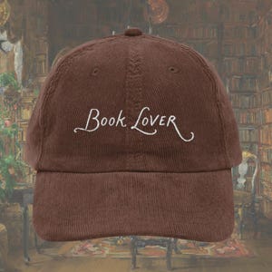 Op de afbeelding: Bruine corduroy baseballcap met de witte geborduurde tekst "Book Lover". De cap heeft een gebogen klep en een knoop aan de bovenkant. De achtergrond toont een bibliotheek.