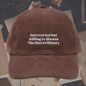 May include: A brown corduroy baseball cap with white embroidered text that reads "introverted but willing to discuss The Secret History".