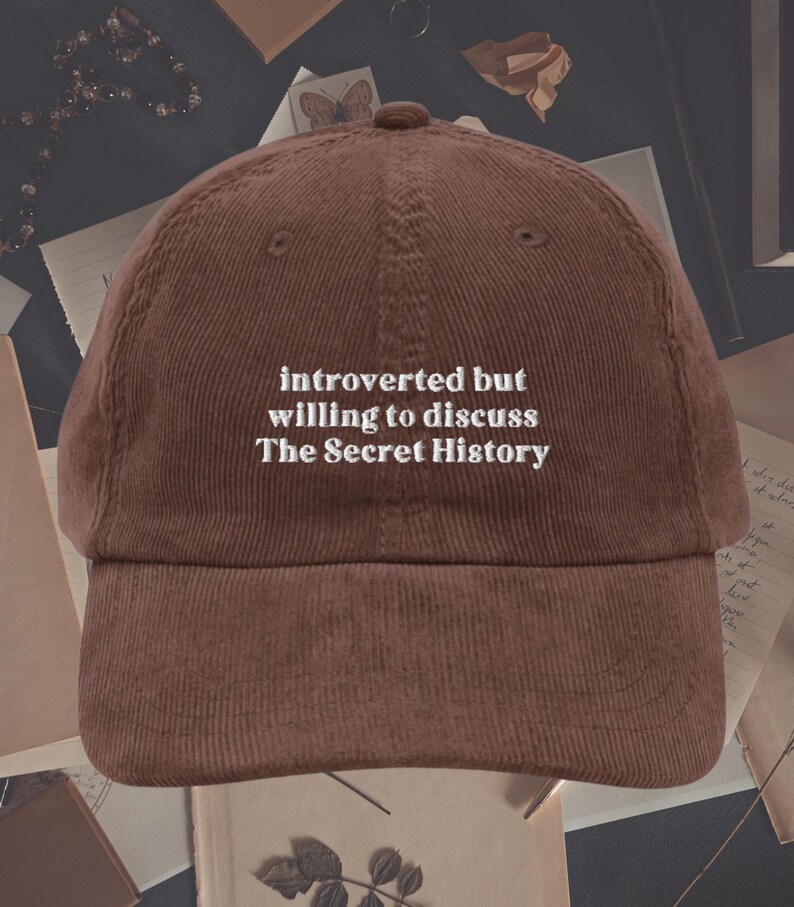 May include: A brown corduroy baseball cap with white embroidered text that reads "introverted but willing to discuss The Secret History".