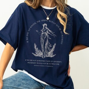 Pu&ograve; includere: Una maglietta oversize blu navy con una stampa grafica bianca di una donna in piedi in un campo di grano. Il testo sulla maglietta recita "Se non avessimo l'inverno, la primavera non sarebbe cos&igrave; piacevole." e "Se non provassimo a volte il sapore dell'avversit&agrave;, la prosperit&agrave; non sarebbe cos&igrave; gradita. Demetra Dea del Raccolto."