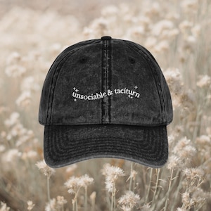 Puede incluir: Gorra de béisbol negra de estilo vintage con las palabras "unsociable & taciturn" bordadas en blanco. La gorra tiene una visera curva y un aspecto desgastado. El fondo es una imagen suave y borrosa de flores blancas.