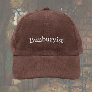 May include: A brown corduroy baseball cap with the word "Bunburyist" embroidered in white. The cap has a curved brim and a rounded crown. The background is a blurred image of a room with bookshelves.