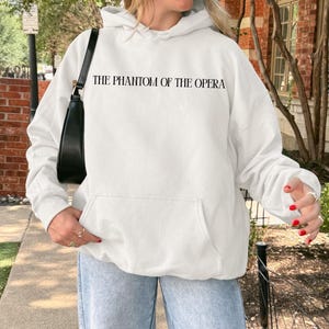 May include: White hooded sweatshirt with the text "THE PHANTOM OF THE OPERA" in black. The sweatshirt has a front pocket and is being worn with light blue jeans. A black purse is hanging from the shoulder.