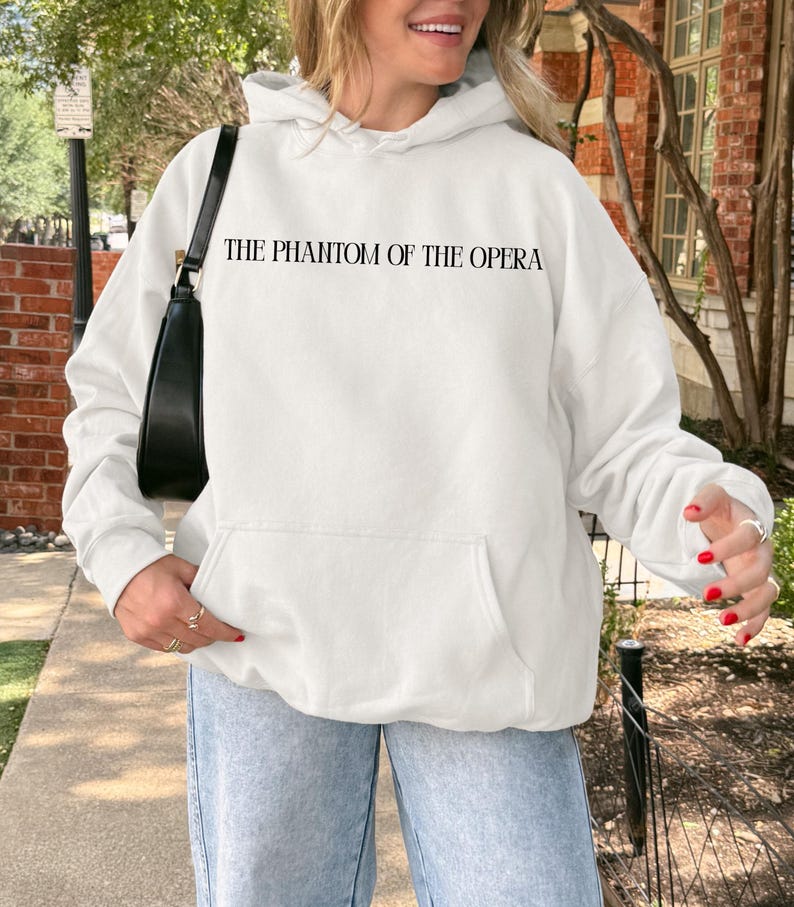 May include: White hooded sweatshirt with the text "THE PHANTOM OF THE OPERA" in black. The sweatshirt has a front pocket and is being worn with light blue jeans. A black purse is hanging from the shoulder.