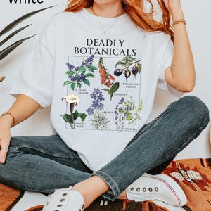 May include: White t-shirt with the text "DEADLY BOTANICALS" and illustrations of various plants. The shirt features images of elderberries, foxglove, and other botanical specimens. The word "white" is in the top left corner of the image.