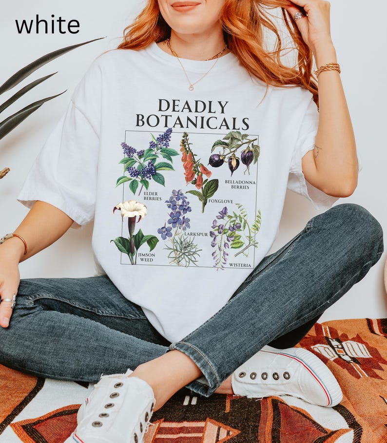 May include: White t-shirt with the text "DEADLY BOTANICALS" and illustrations of various plants. The shirt features images of elderberries, foxglove, and other botanical specimens. The word "white" is in the top left corner of the image.