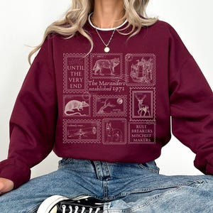 K&ouml;nnte beinhalten: Burgunderfarbener Pullover mit einem grafischen Druck von Vintage-Briefmarken mit einem Wolf, einem Schloss, einer Ratte, einem Baum, einem Reh, einem Teleskop und einem Hund. Der Text auf den Briefmarken lautet "The Marauders established 1971" und "Rule Breakers Mischief Makers".