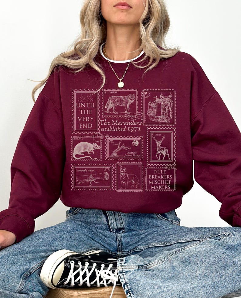 K&ouml;nnte beinhalten: Burgunderfarbener Pullover mit einem grafischen Druck von Vintage-Briefmarken mit einem Wolf, einem Schloss, einer Ratte, einem Baum, einem Reh, einem Teleskop und einem Hund. Der Text auf den Briefmarken lautet "The Marauders established 1971" und "Rule Breakers Mischief Makers".