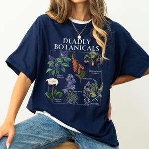 May include: Navy blue t-shirt with a white collar featuring a botanical illustration. The graphic includes the text "DEADLY BOTANICALS" and illustrations of various plants, including elderberries, foxglove, and belladonna berries. The word "navy" is printed at the bottom.