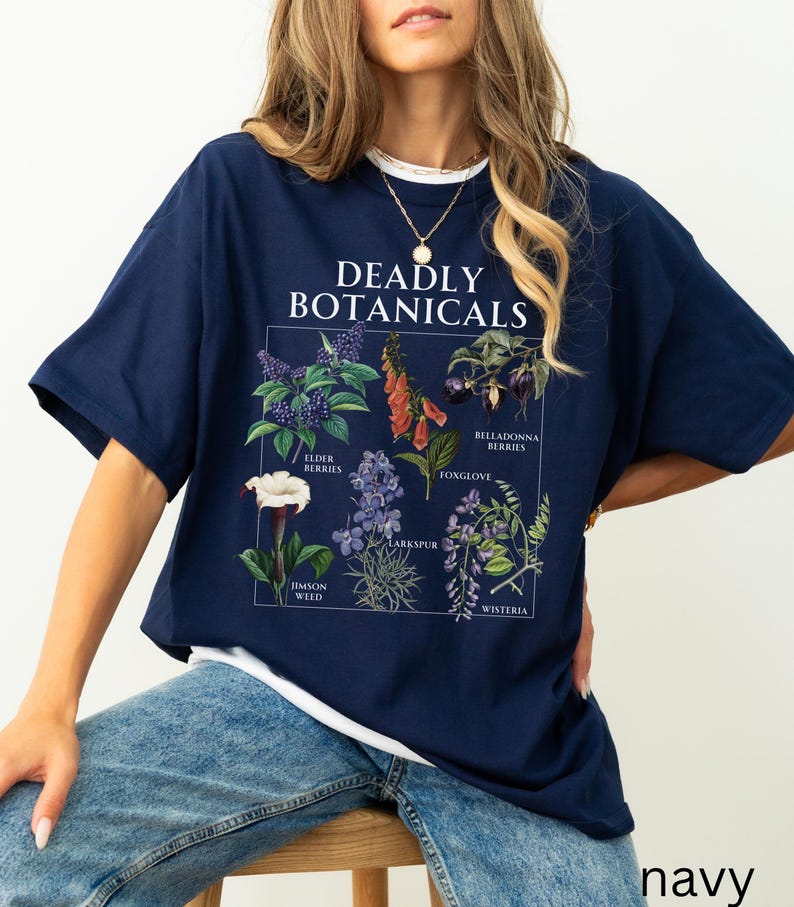 May include: Navy blue t-shirt with a white collar featuring a botanical illustration. The graphic includes the text "DEADLY BOTANICALS" and illustrations of various plants, including elderberries, foxglove, and belladonna berries. The word "navy" is printed at the bottom.
