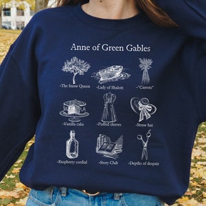 Op de afbeelding: Een donkerblauw sweatshirt met een witte afbeelding van illustraties en tekst die betrekking hebben op het boek "Anne of Green Gables". De tekst omvat "The Snow Queen", "Lady of Shalott", "Carrots", "Vanilla cake", "Puffed sleeves", "Straw hat", "Raspberry cordial", "Story Club" en "Depths of despair".