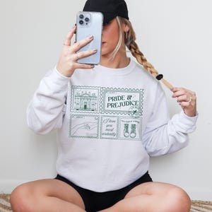 Op de afbeelding: Witte sweatshirt met een groene postzegelontwerp. De afbeelding bevat de woorden "Pride & Prejudice" en illustraties van een gebouw, handen en laarzen, samen met de zin "I love you most ardently."