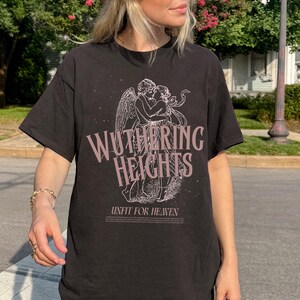 May include: Black t-shirt featuring the text "Wuthering Heights" in a stylized font, with an illustration of two figures embracing. Below the text is the phrase "Unfit For Heaven". The shirt is a dark color.