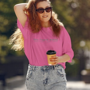 May include: A woman wearing a pink t-shirt with the text "Very Fond Of Walking" and blue jeans is holding a brown paper cup of coffee.