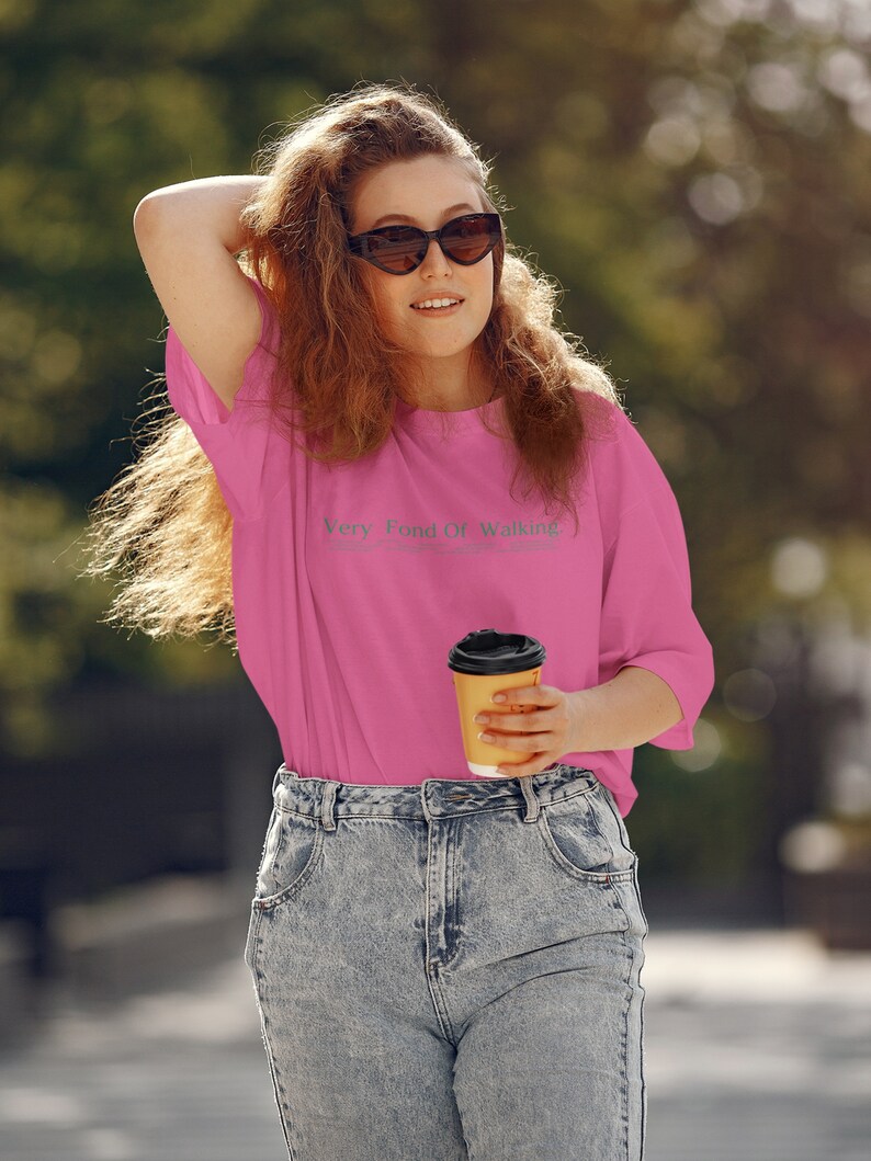 May include: A woman wearing a pink t-shirt with the text "Very Fond Of Walking" and blue jeans is holding a brown paper cup of coffee.