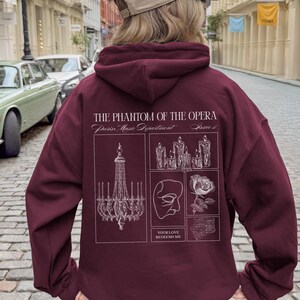 May include: Burgundy hoodie featuring a graphic design from "The Phantom of the Opera." The design includes a chandelier, candles, a mask, and a rose, with the text "YOUR LOVE REDEEMS ME."