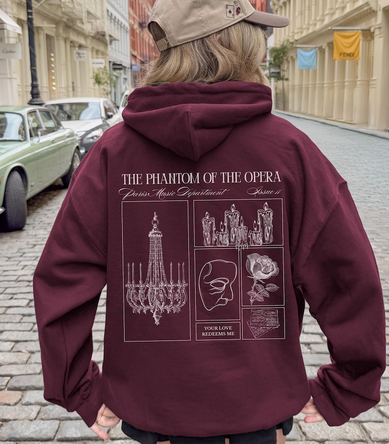 May include: Burgundy hoodie featuring a graphic design from "The Phantom of the Opera." The design includes a chandelier, candles, a mask, and a rose, with the text "YOUR LOVE REDEEMS ME."