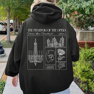 May include: Black hoodie featuring a graphic design from "The Phantom of the Opera." The design includes a chandelier, candles, a mask, a rose, and sheet music. The text "THE PHANTOM OF THE OPERA" and "YOUR LOVE REDEEMS ME" are also present.