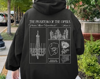 May include: Black hoodie featuring a graphic design from "The Phantom of the Opera." The design includes a chandelier, candles, a mask, a rose, and sheet music. The text "THE PHANTOM OF THE OPERA" and "YOUR LOVE REDEEMS ME" are also present.