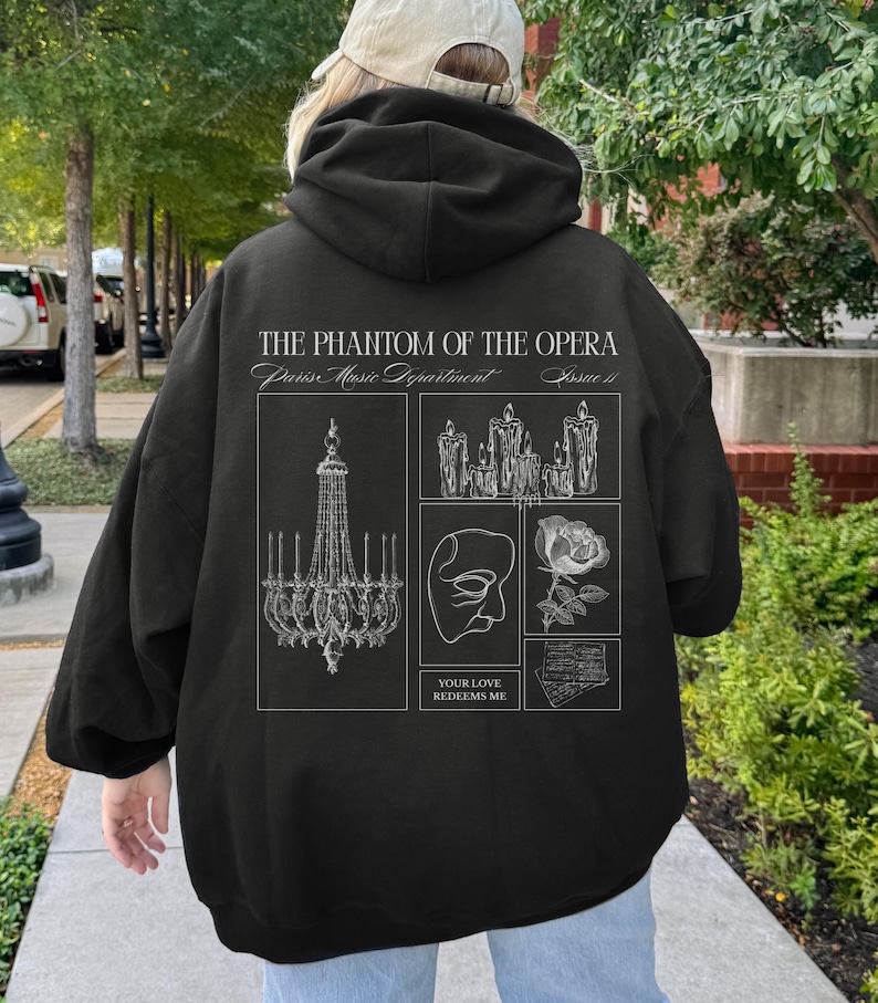 May include: Black hoodie featuring a graphic design from "The Phantom of the Opera." The design includes a chandelier, candles, a mask, a rose, and sheet music. The text "THE PHANTOM OF THE OPERA" and "YOUR LOVE REDEEMS ME" are also present.
