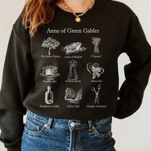 Op de afbeelding: Zwart sweatshirt met een witte grafische print van illustraties en tekst die "Anne of Green Gables" zegt. De illustraties omvatten een boom, een boot, wortels, een taart, een jurk, een hoed, een fles, een boek en een schaar. Elke illustratie heeft tekst eronder.