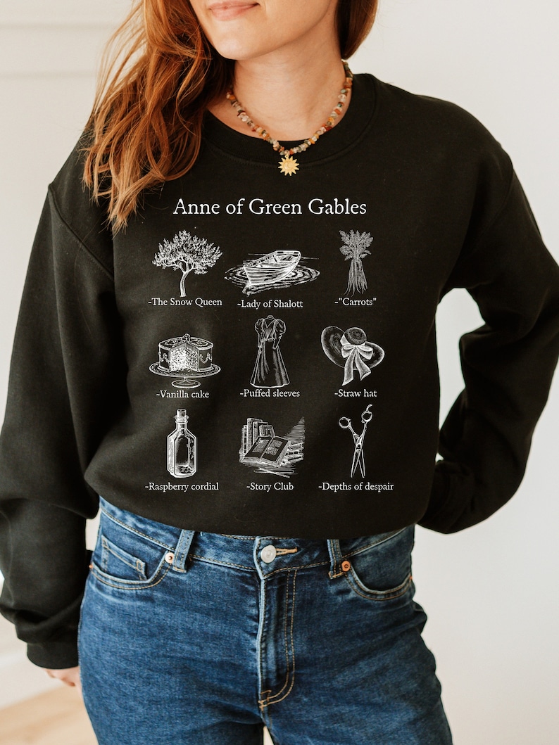 Op de afbeelding: Zwart sweatshirt met een witte grafische print van illustraties en tekst die "Anne of Green Gables" zegt. De illustraties omvatten een boom, een boot, wortels, een taart, een jurk, een hoed, een fles, een boek en een schaar. Elke illustratie heeft tekst eronder.