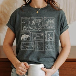 May include: A dark gray t-shirt with a white graphic print of stamps featuring illustrations of a wolf, a castle, a rat, a tree, a deer, a lizard, a dog, and text that reads "Until the very end. The Marauders established 1971. Rule Breakers Mischief Makers."
