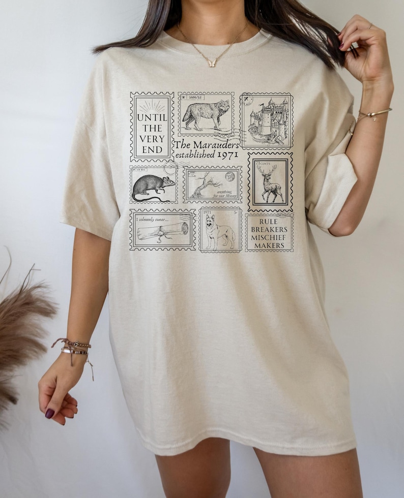 May include: A beige t-shirt with a graphic print of stamps featuring illustrations of a wolf, a castle, a rat, a tree, a deer, a dog, and a telescope. The text on the stamps reads "The Marauders established 1971", "Until the very end", "I solemnly swear", and "Rule breakers mischief makers".